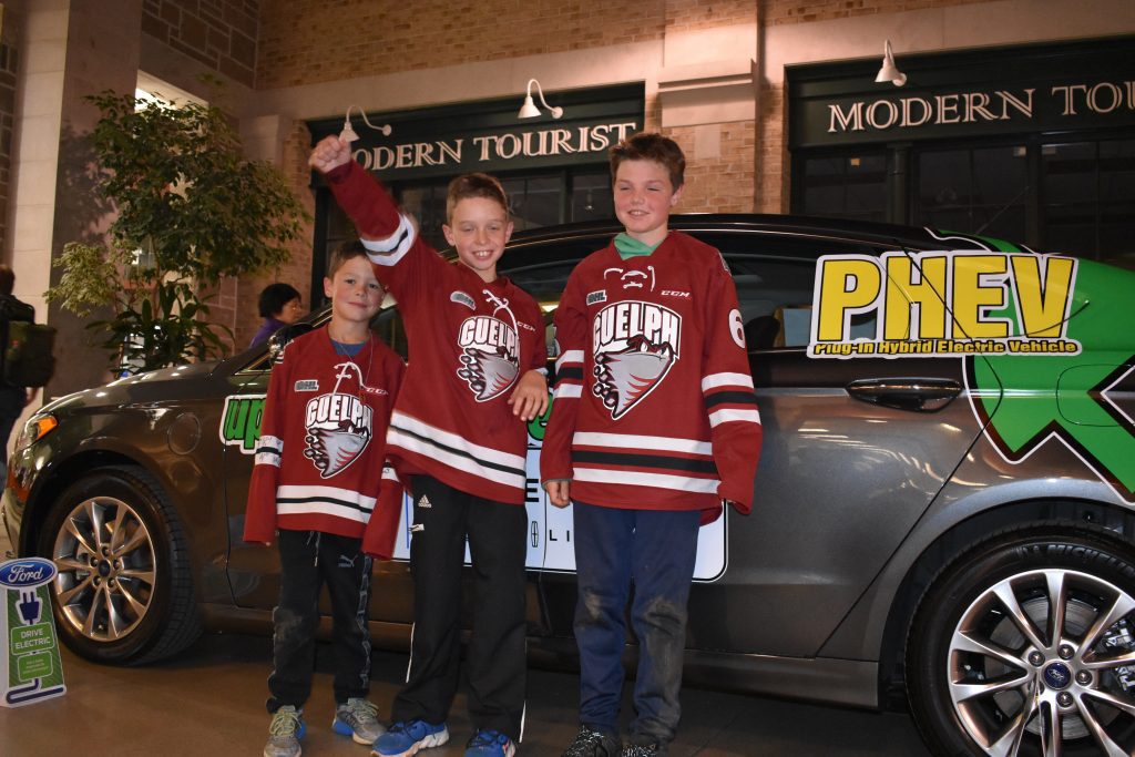 Three young boys in hockey jerseys standing in front of a hybrid electric car