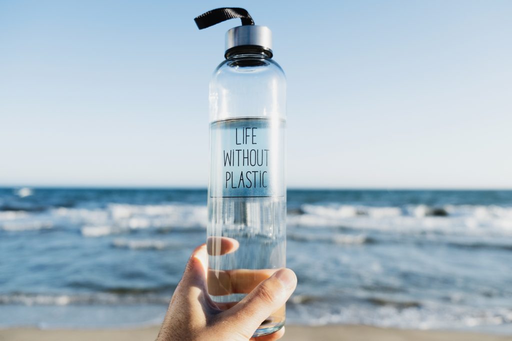 closeup of a caucasian man holding a glass reusable water bottle with the text life without plastic written in it, on the beach, with the ocean in the background