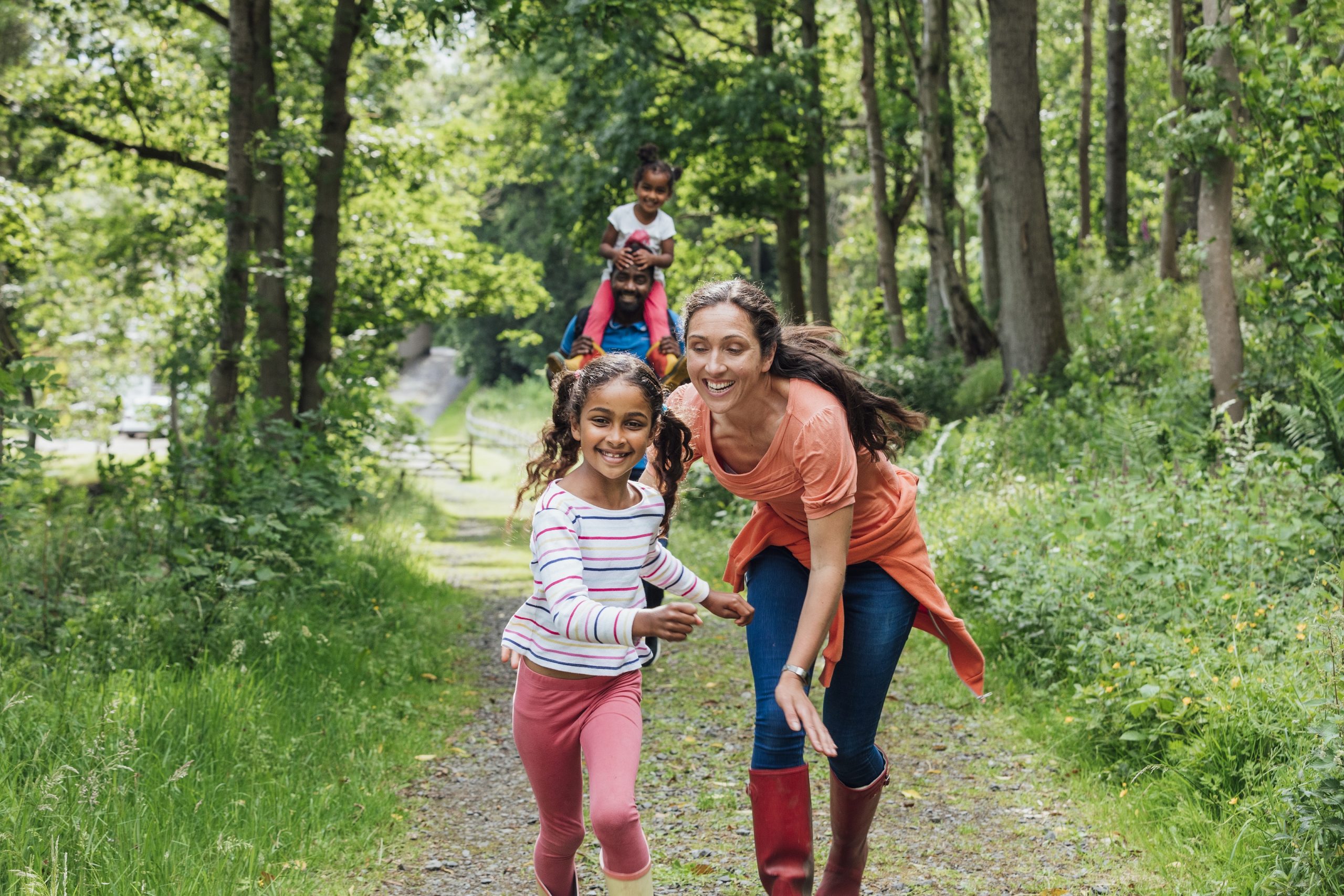 Happy family running up a footpath through the woods in their wellies in Northumberland. One of the children is on her father's shoulders.