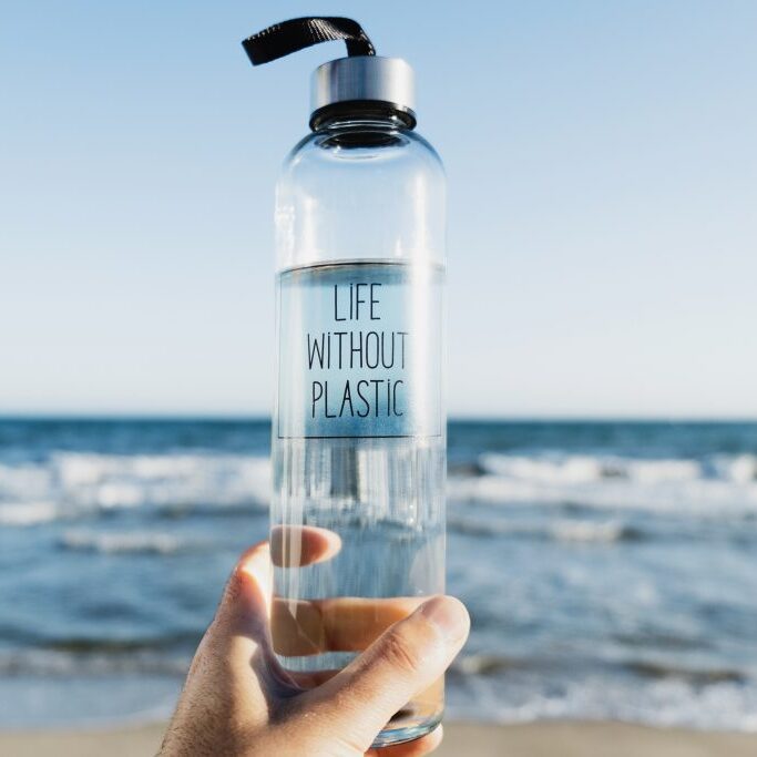 closeup of a caucasian man holding a glass reusable water bottle with the text life without plastic written in it, on the beach, with the ocean in the background